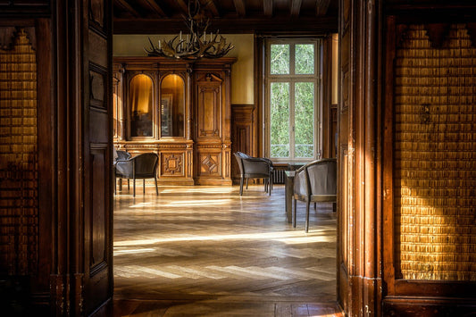 Sunlit antique sitting room with ornate 19th-century wooden cabinetry, parquet flooring, and vintage seating near large windows